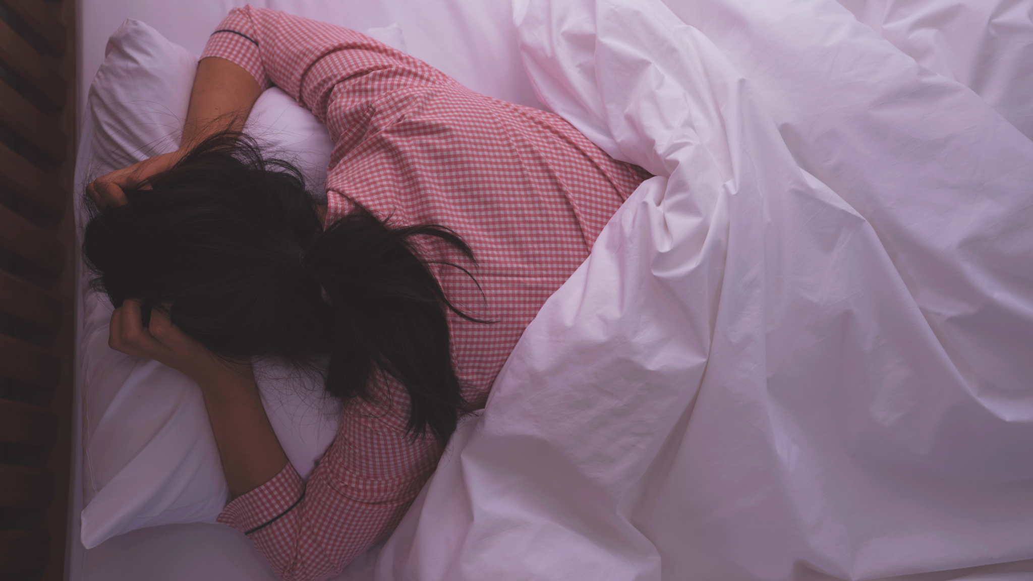 Woman in pink pajamas lying on bed with white sheets, struggling to sleep, representing hormonal sleep disruptions and restful sleep challenges.