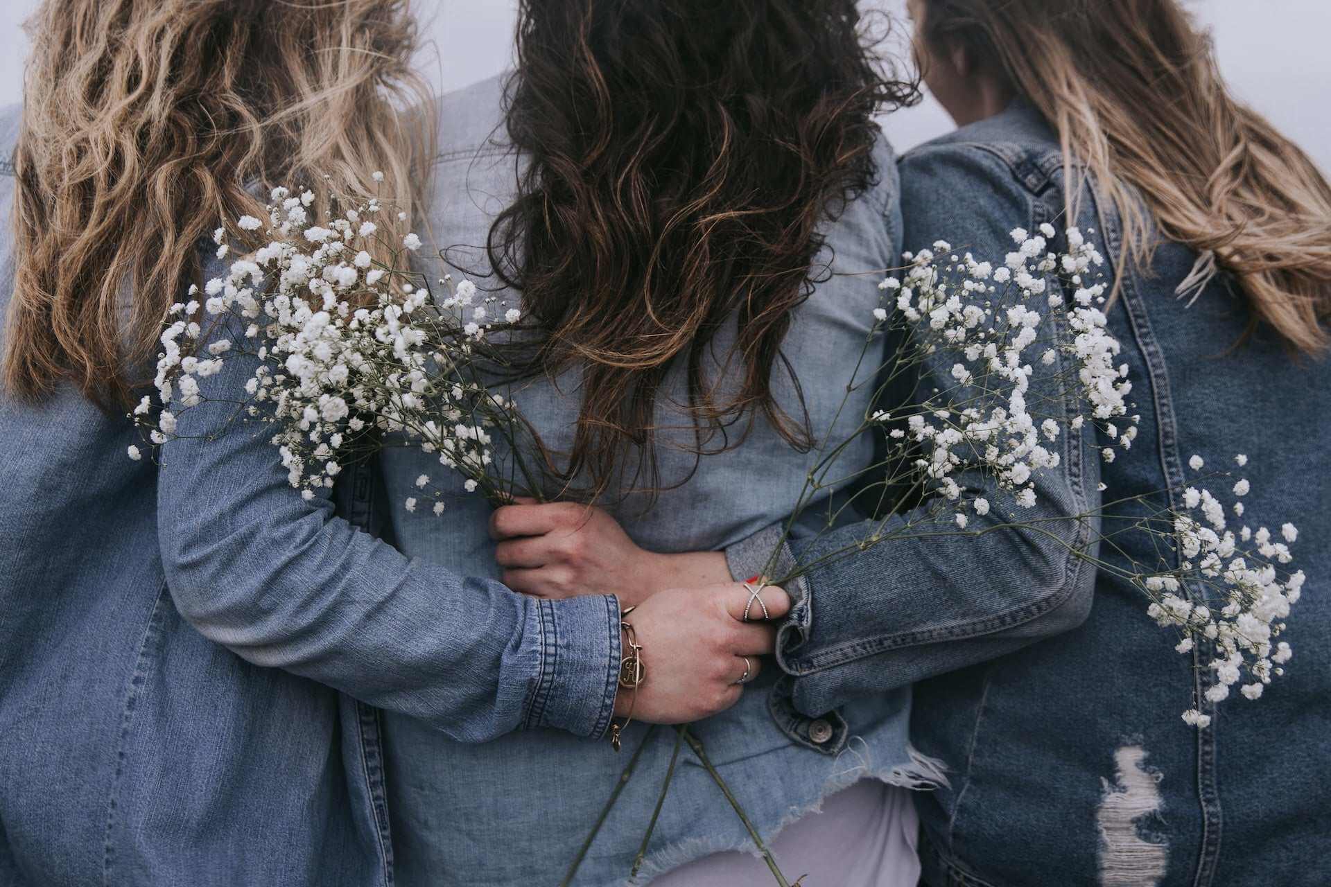 three women in denim jackets holding white flowers from behind representing support and awareness for hypertonic pelvic floor muscles condition